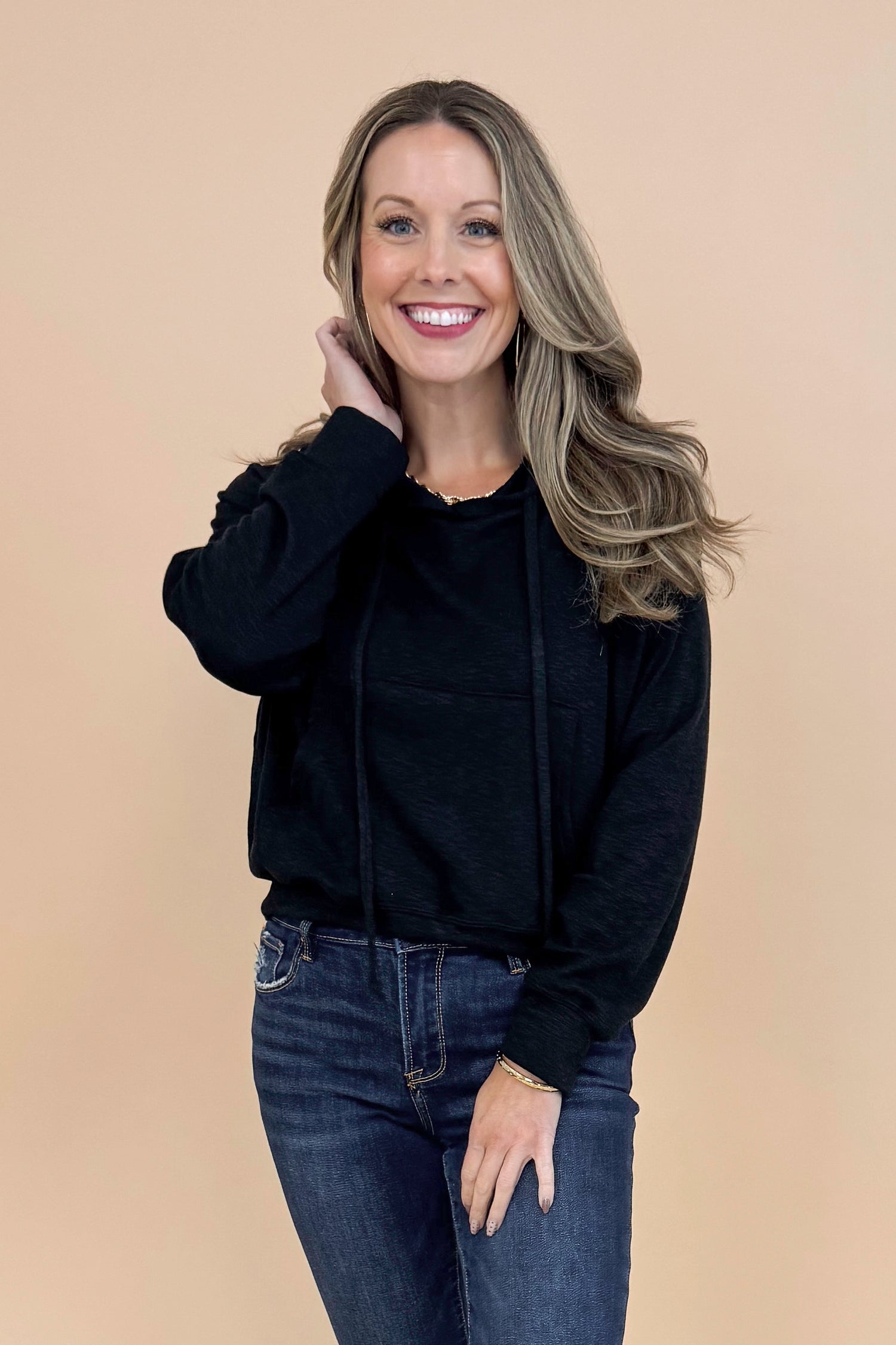 Woman wearing Black Hooded Sweatshirt and jeans with a tan background. 