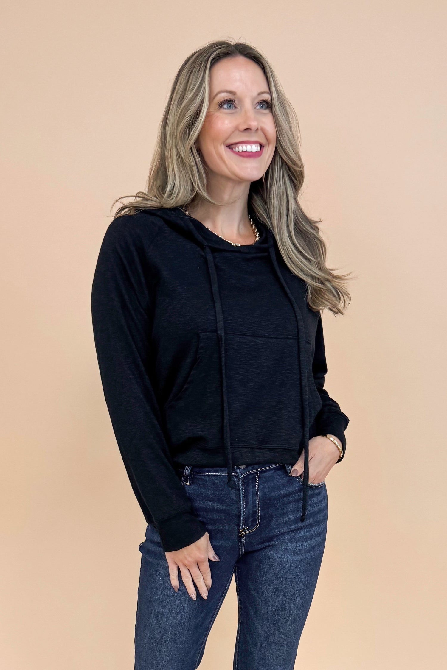 Woman wearing Black Hooded Sweatshirt and jeans with a tan background. 