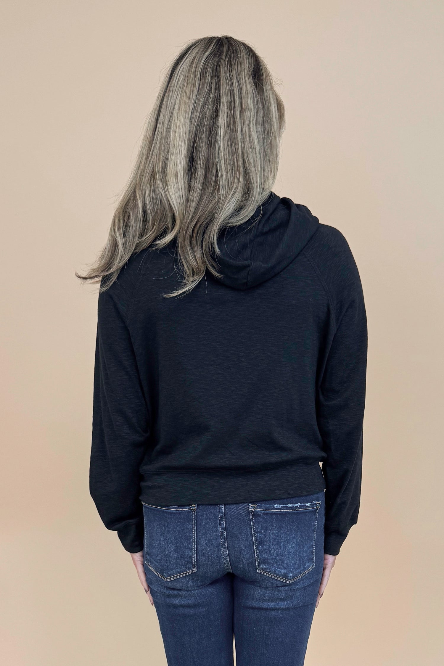 Back view of woman wearing Black Hooded Sweatshirt and jeans with a tan background. 