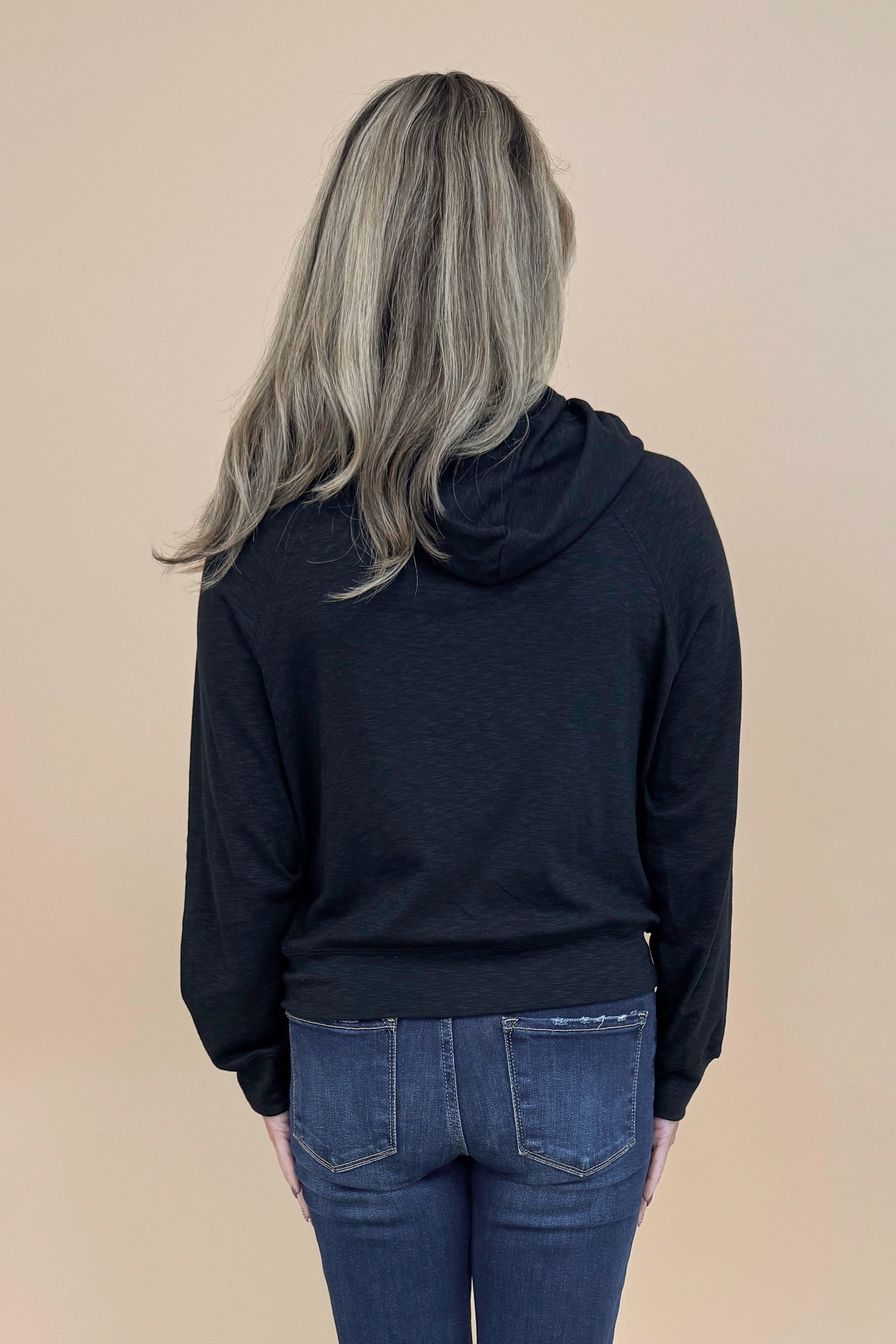 Back view of woman wearing Black Hooded Sweatshirt and jeans with a tan background. 