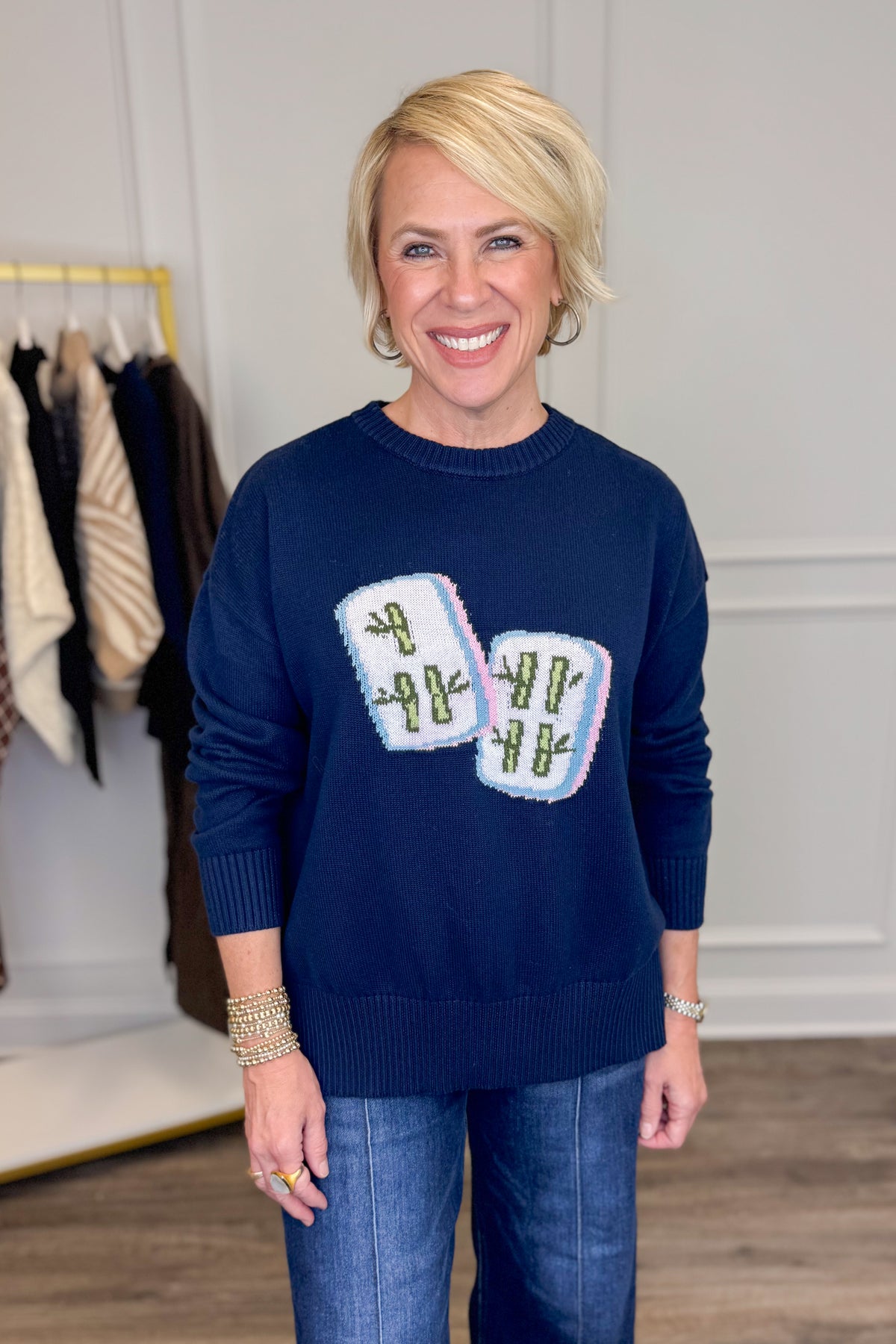 A smiling blonde woman wearing a navy blue crewneck sweater with two white mahjong tiles on the front, paired with blue jeans, standing in an indoor boutique setting.