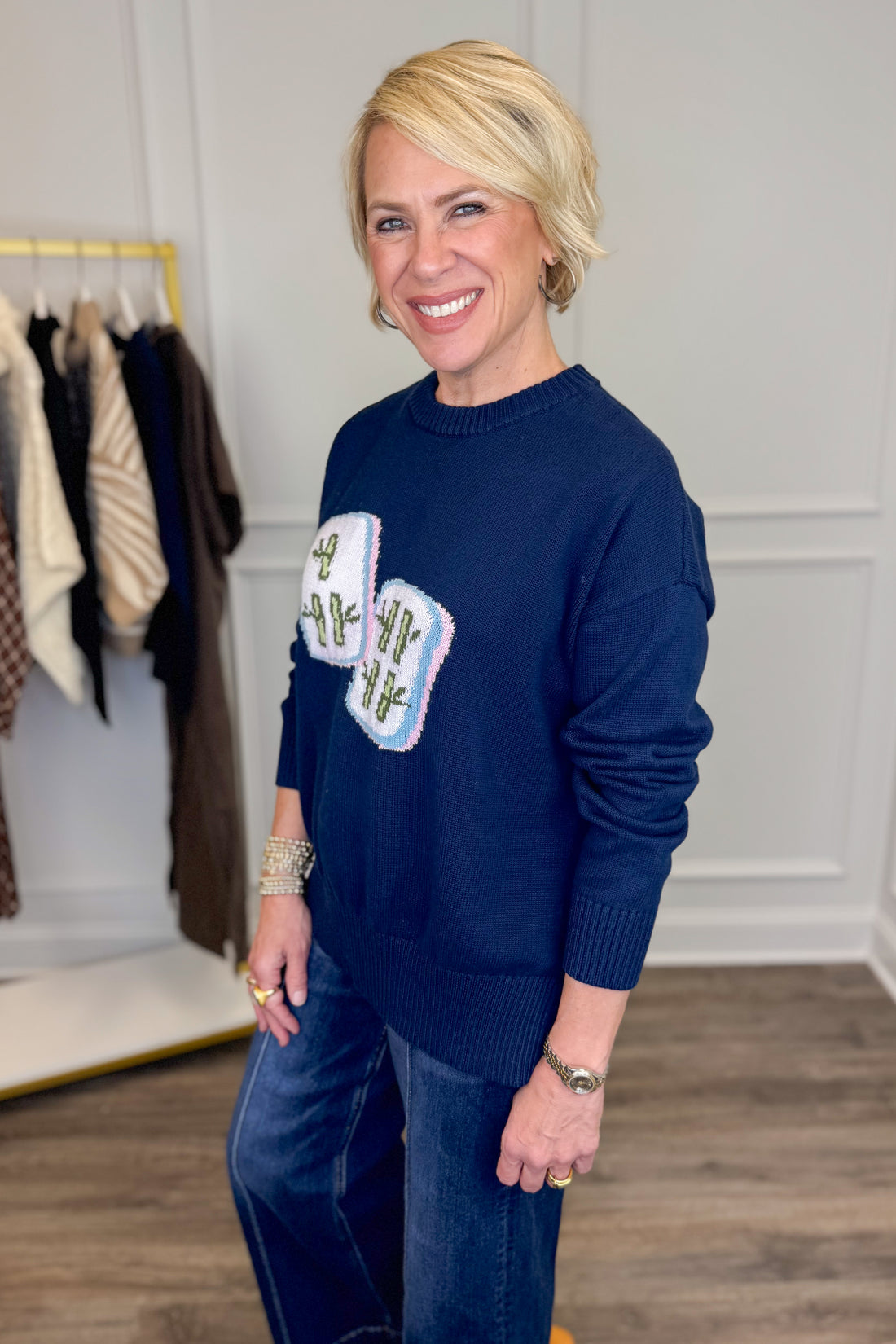 A smiling blonde woman wearing a navy blue crewneck sweater with two white mahjong tiles on the front, paired with blue jeans, standing in an indoor boutique setting.