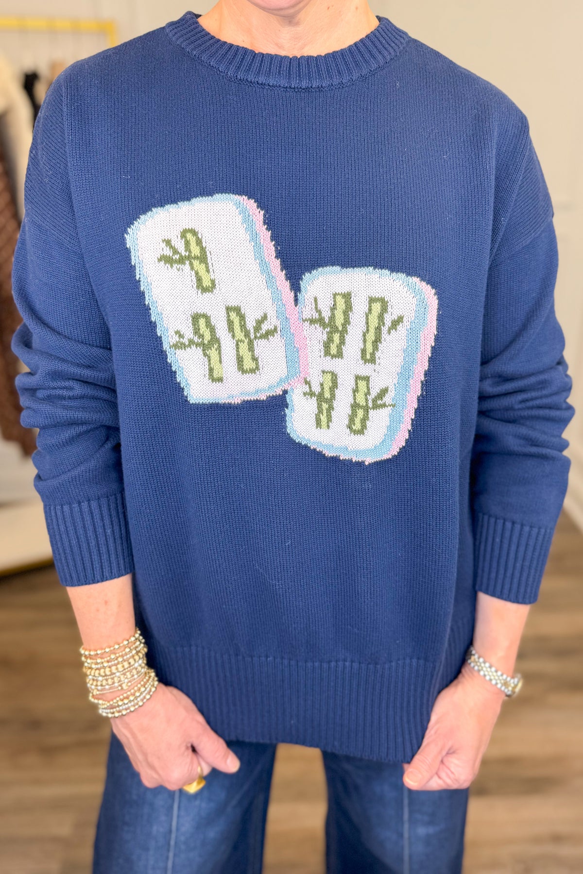 A smiling blonde woman wearing a navy blue crewneck sweater with two white mahjong tiles on the front, paired with blue jeans, standing in an indoor boutique setting.