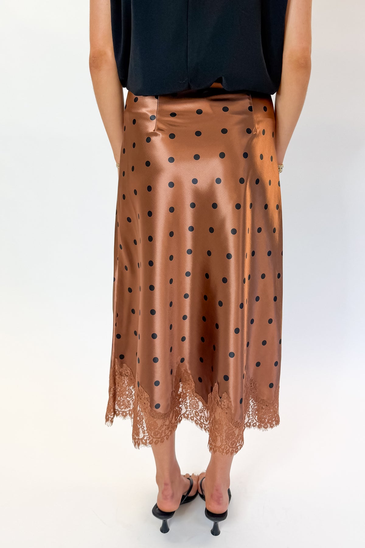 Back detail view of woman wearing Swagger Boutique’s Camel Polka Dot Satin Skirt with black top against white studio background.