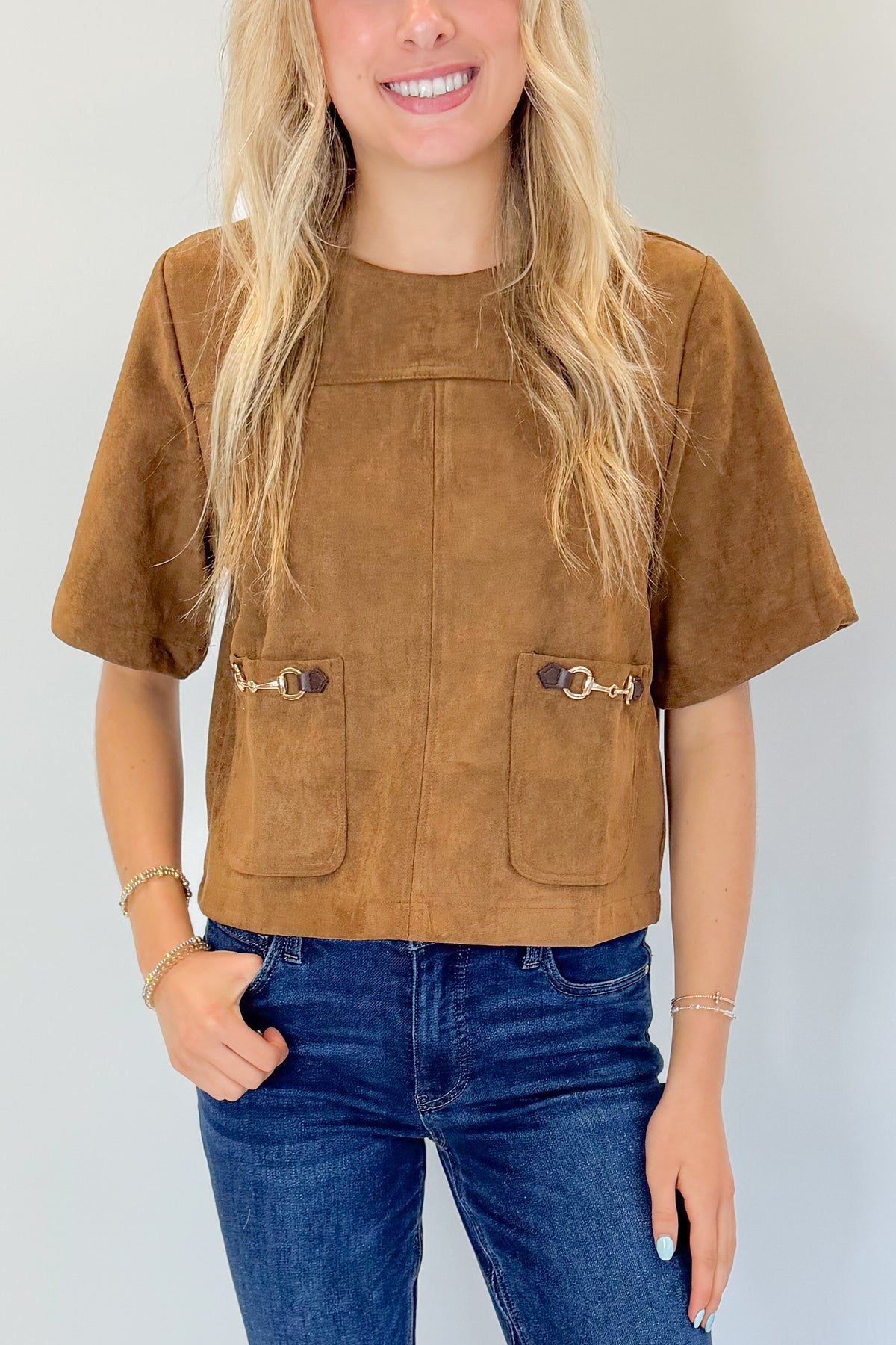 Front detail view of woman wearing Swagger Boutique’s Camel Suede Horsebit Pocket Top by Entro with jeans against white studio background.