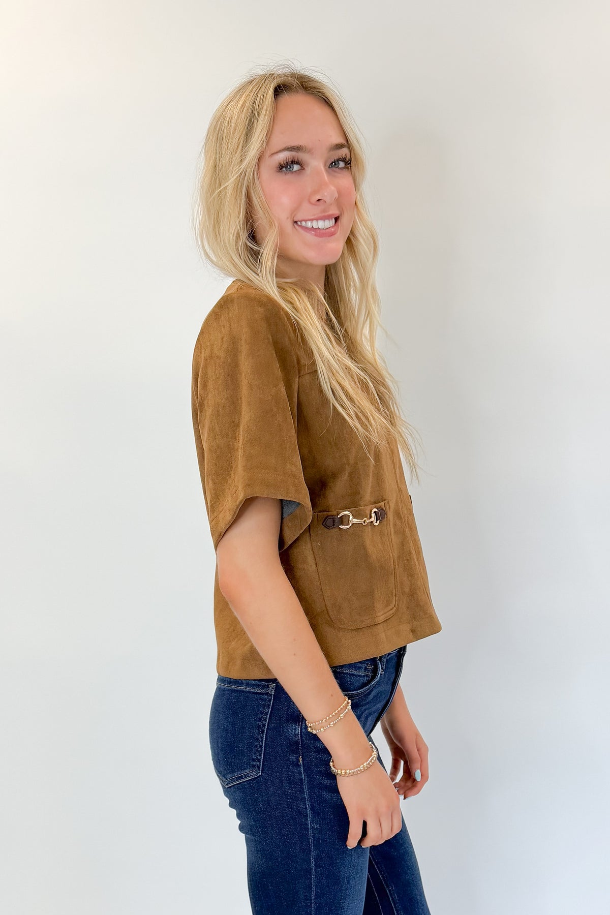Side view of woman wearing Swagger Boutique’s Camel Suede Horsebit Pocket Top by Entro with jeans against white studio background.