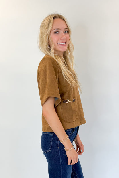 Side view of woman wearing Swagger Boutique’s Camel Suede Horsebit Pocket Top by Entro with jeans against white studio background.