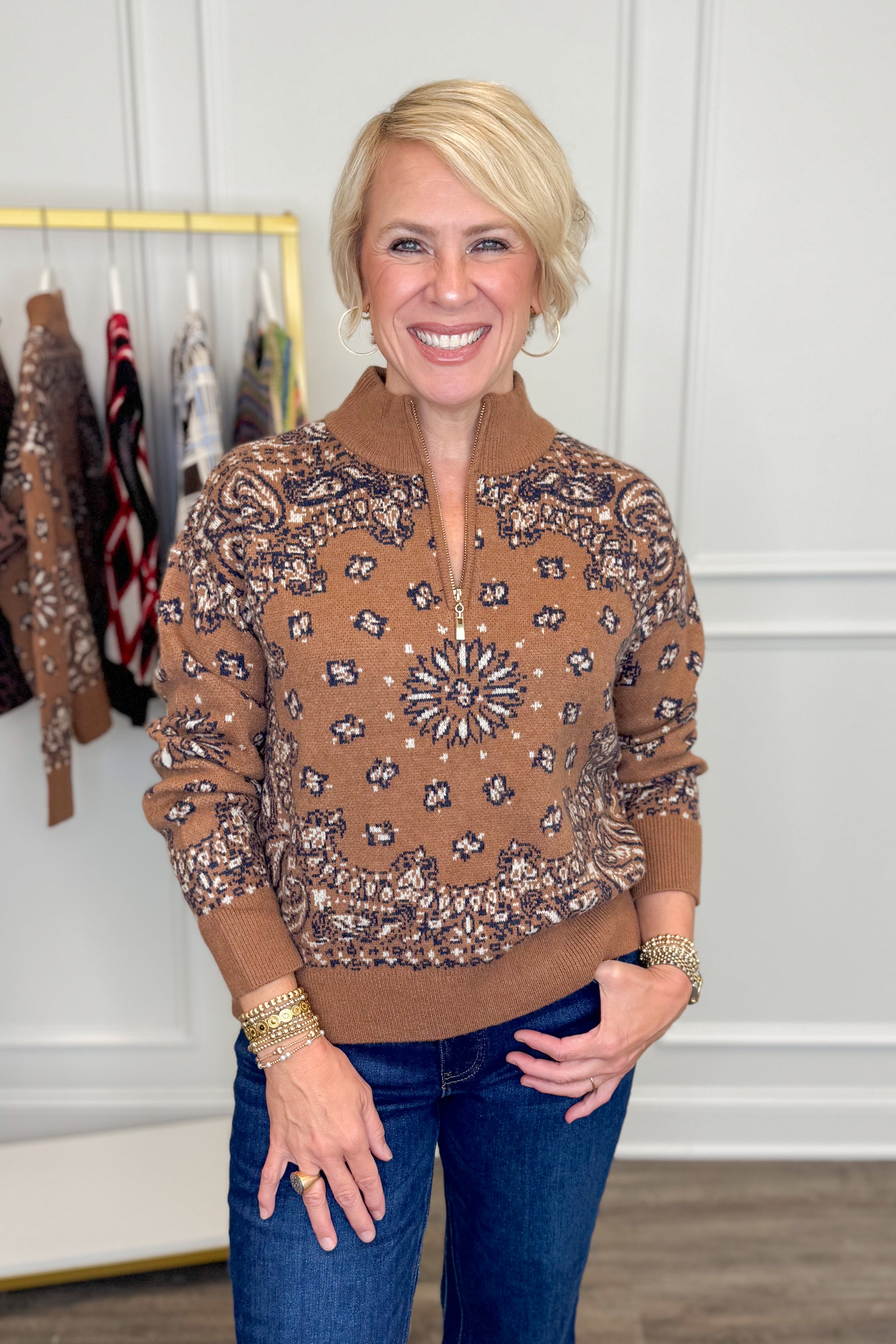 Front view of woman wearing Swagger Boutique’s Camel Bandana Print Half Zip Sweater with jeans with clothing hanging in the background.