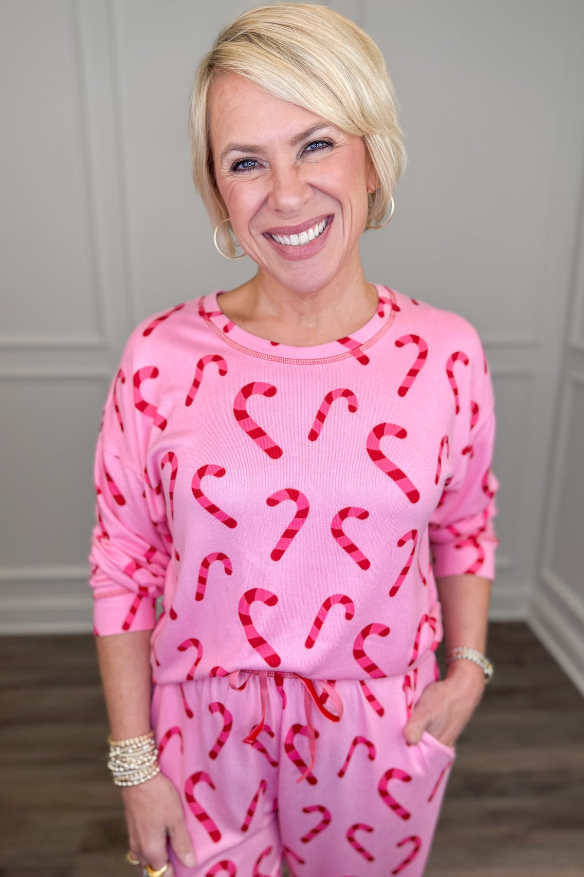 A smiling blonde woman wearing a pink long-sleeve pajama top and matching pants with a red candy cane pattern, standing indoors against a light gray paneled wall.