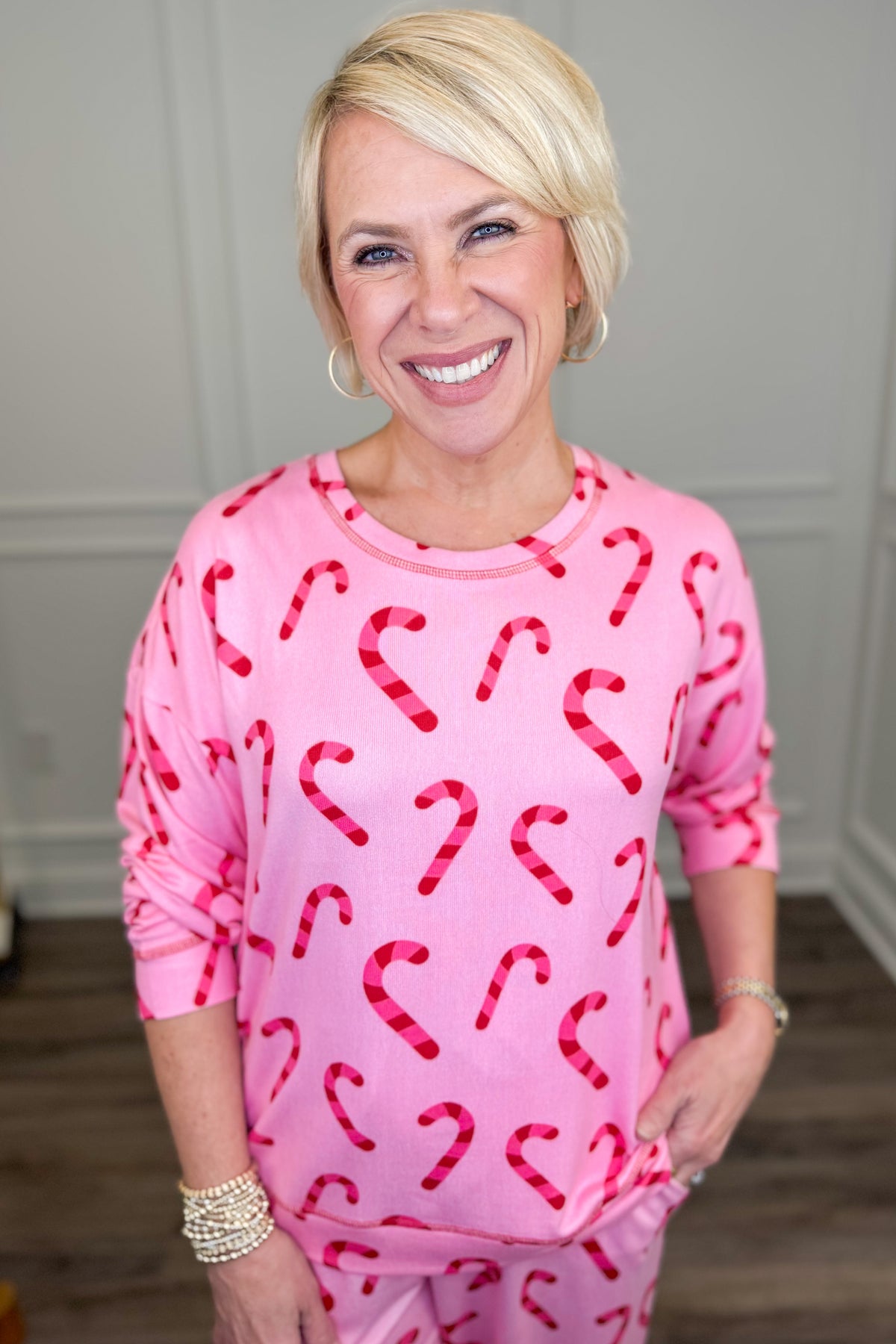 A smiling blonde woman wearing a pink long-sleeve top and matching pants with a red and white candy cane pattern, standing in an indoor room with light gray walls and wood flooring.