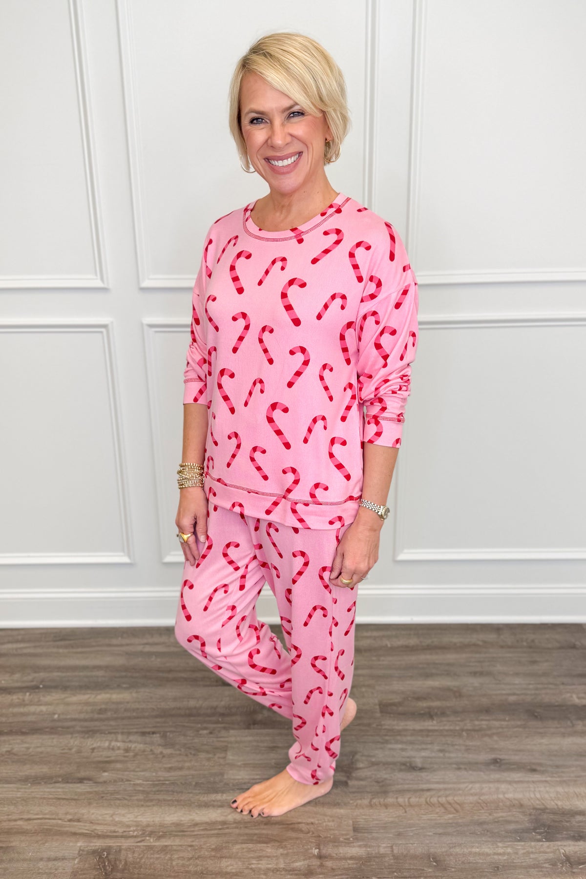 A smiling blonde woman stands barefoot indoors wearing a pink pajama set with a red candy cane pattern. The set includes a long-sleeve top and matching pants, photographed against a white paneled wall and wood floor.