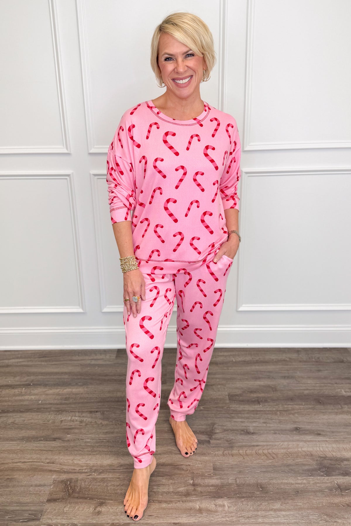 A woman modeling a pink two-piece pajama set with a red candy cane pattern, standing on a wood floor against a white paneled wall.