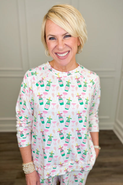 Model wearing the Champagne Toast Annie Pajama Set, featuring a playful champagne bottle and cocktail glass print in pink, green, and gold on a white background. The set includes a relaxed-fit, long-sleeve crewneck top and matching pajama shorts with side pockets.