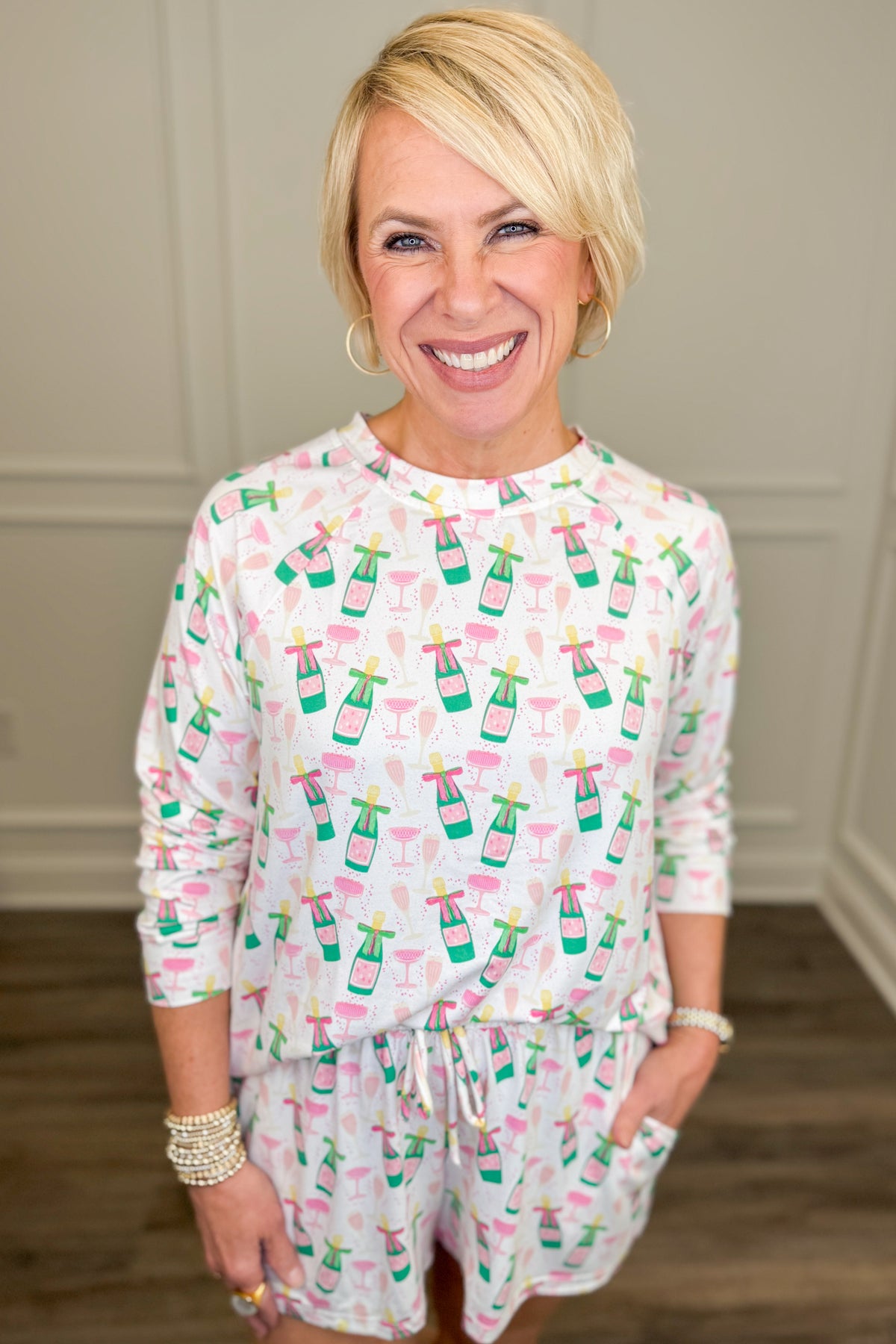 Model wearing the Champagne Toast Annie Pajama Set, featuring a playful champagne bottle and cocktail glass print in pink, green, and gold on a white background. The set includes a relaxed-fit, long-sleeve crewneck top and matching pajama shorts with side pockets.