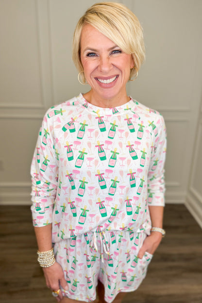 Model wearing the Champagne Toast Annie Pajama Set, featuring a playful champagne bottle and cocktail glass print in pink, green, and gold on a white background. The set includes a relaxed-fit, long-sleeve crewneck top and matching pajama shorts with side pockets.