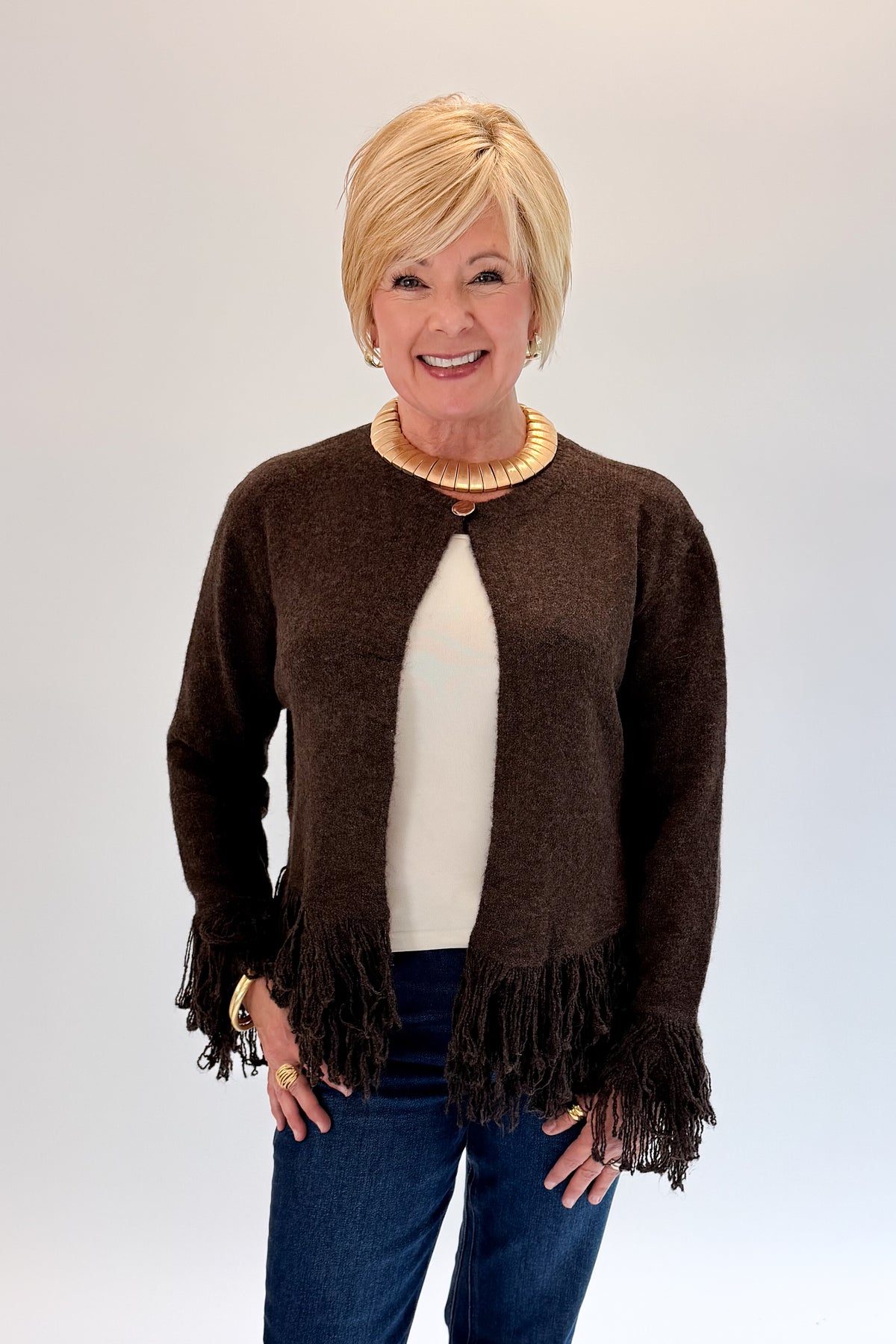 A smiling blonde woman wearing a brown open-front knit cardigan with fringe trim, layered over a cream top and blue jeans, standing against a light gray studio background.
