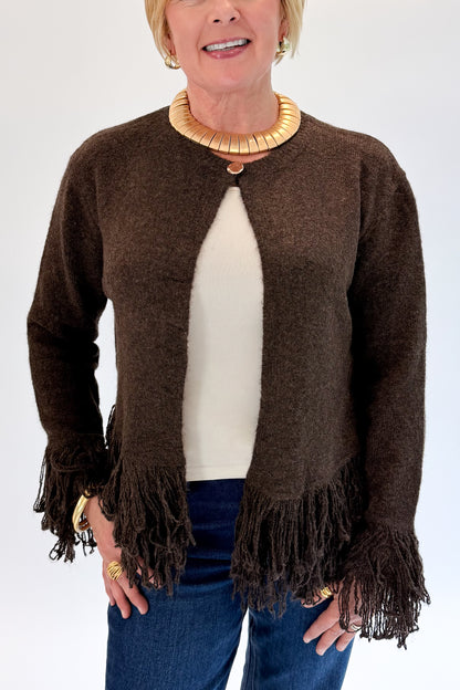 A woman wearing a dark brown open-front knit cardigan with fringe trim at the hem and cuffs, layered over a cream ribbed top and blue jeans, against a plain white studio background.