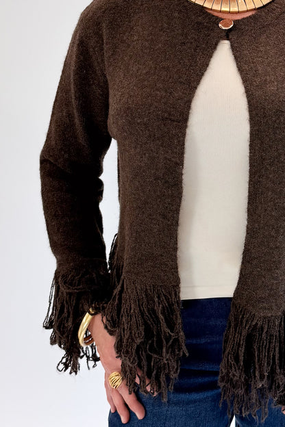 A woman wearing a brown open-front knit cardigan with fringe trim, layered over a cream top and blue jeans, standing against a light gray studio background.