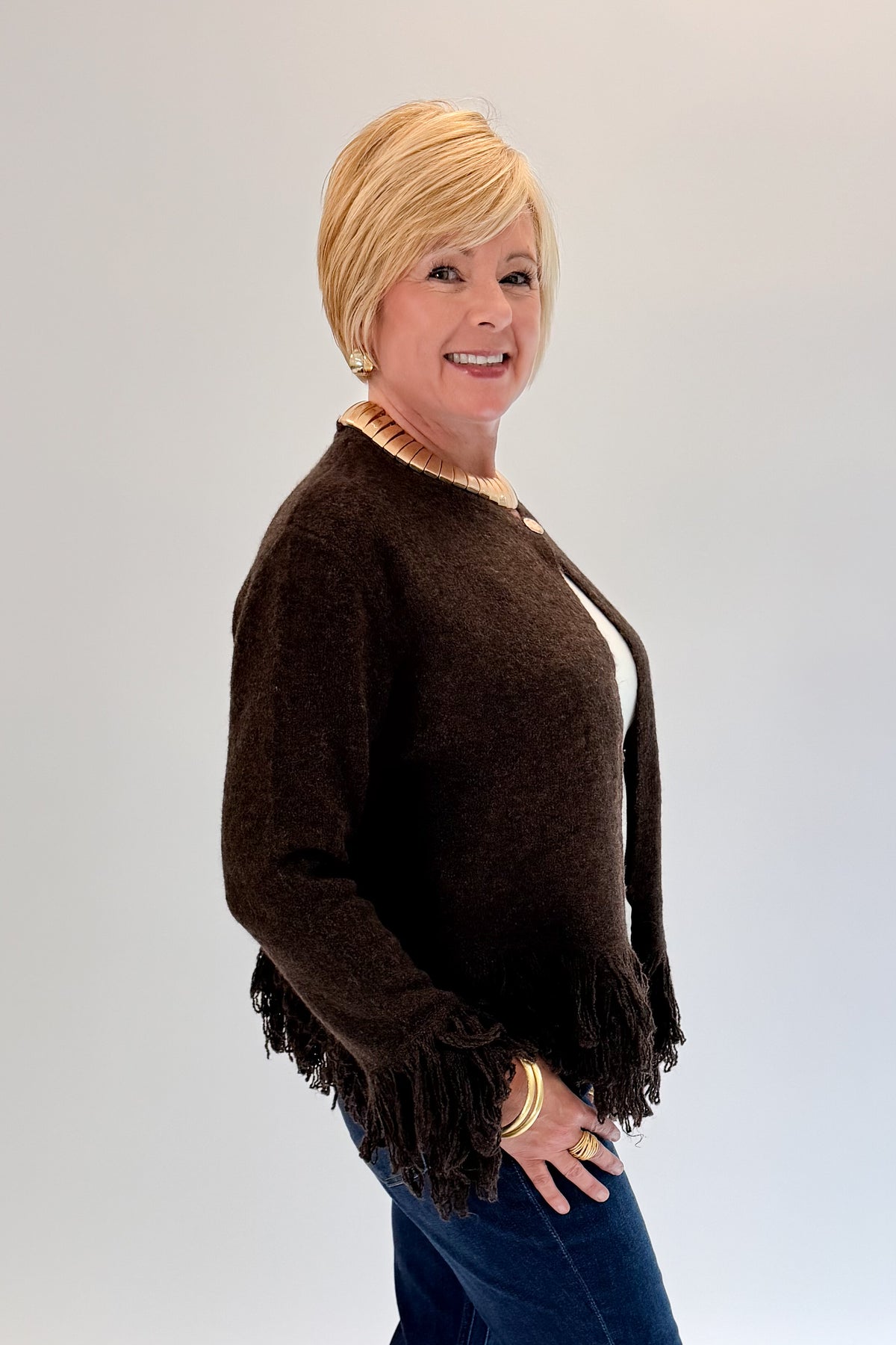 A woman with short blonde hair wearing a dark brown fringed cardigan over a white top and blue jeans, posing against a light gray studio background.