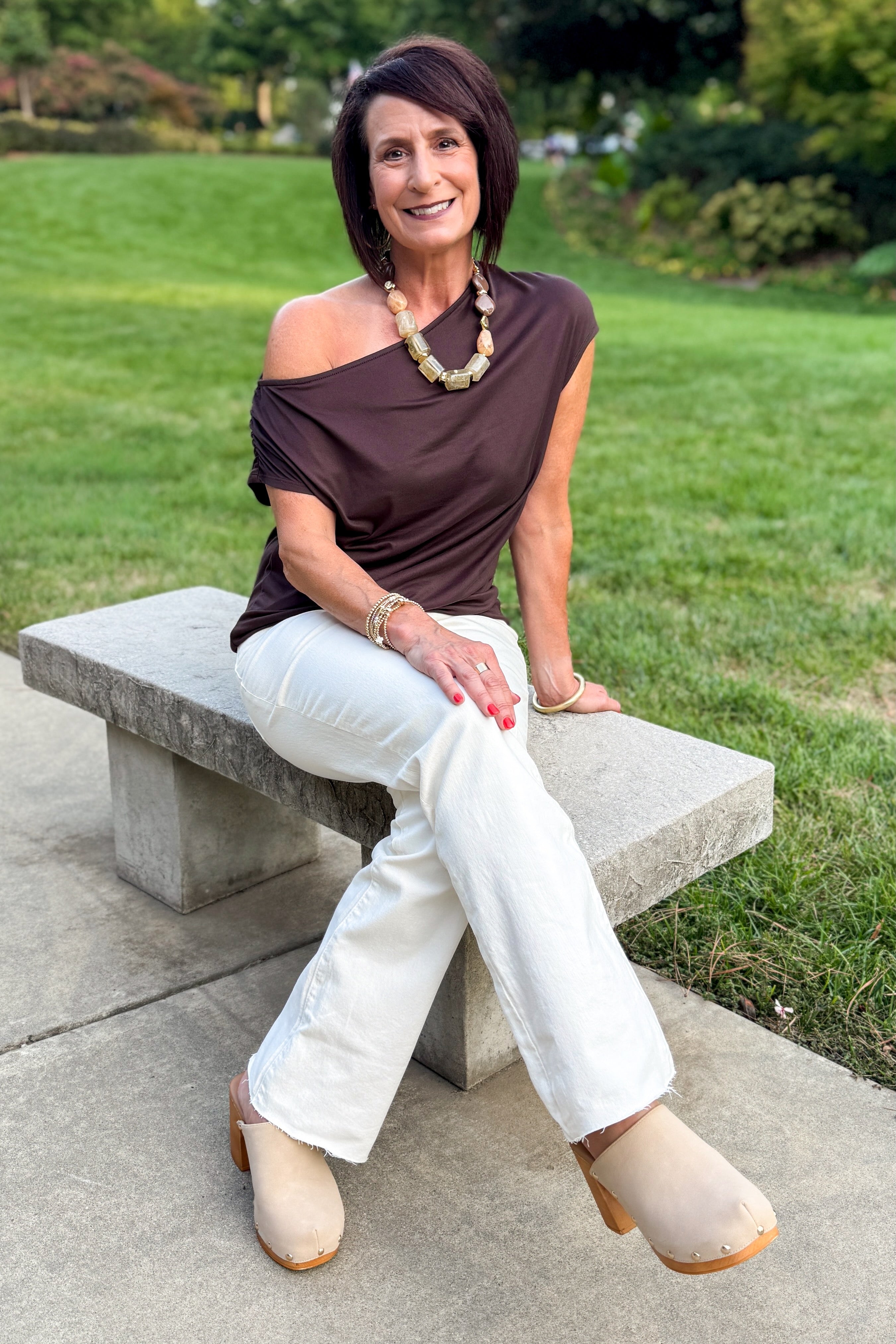 Front view of woman wearing Swagger Boutique’s Chocolate Brown Off The Shoulder Top with jeans sitting on a bench outside.