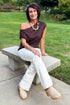 Front view of woman wearing Swagger Boutique’s Chocolate Brown Off The Shoulder Top with jeans sitting on a bench outside.
