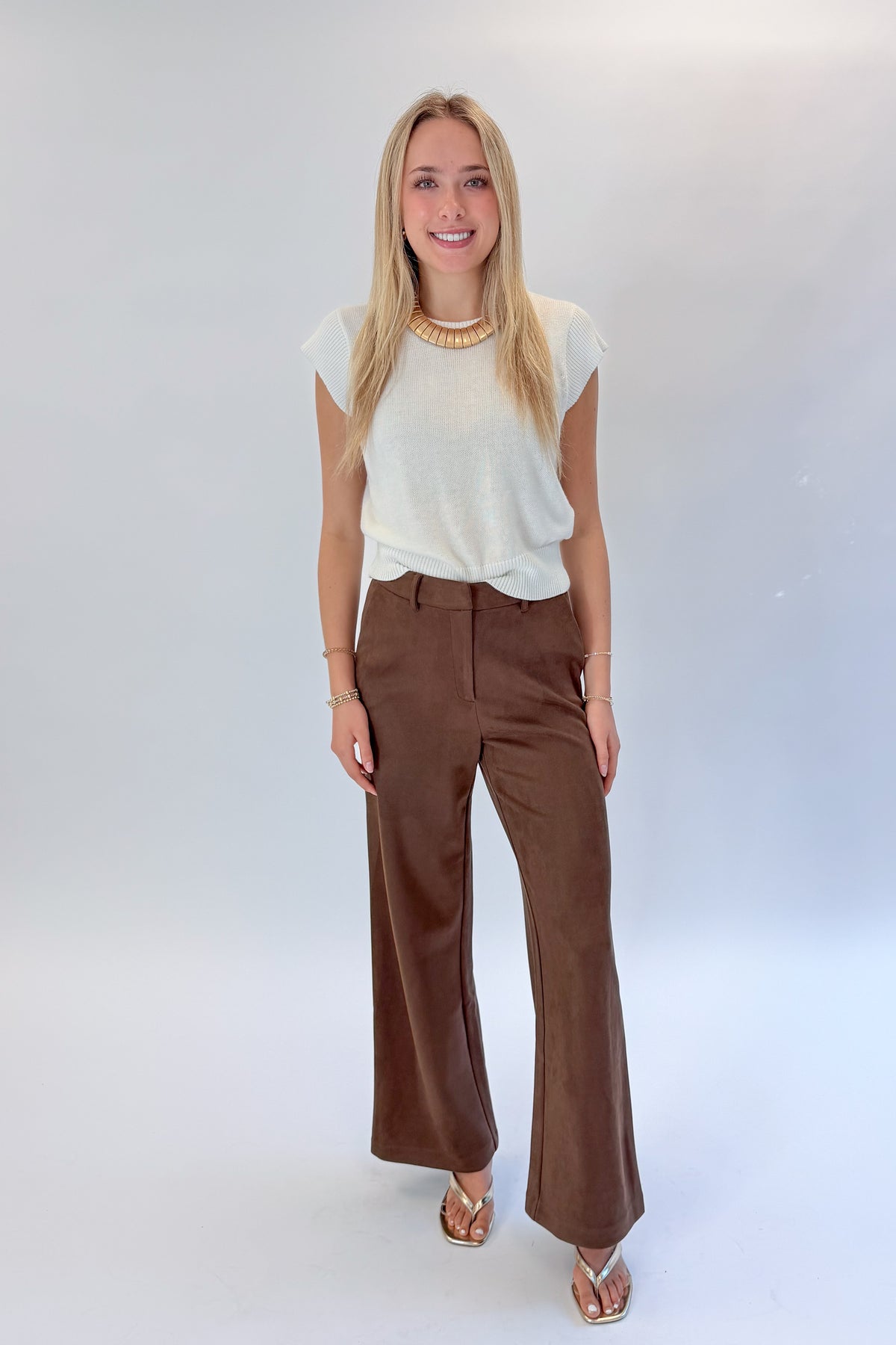 Front view of woman wearing Swagger Boutique’s Chocolate Brown Suede Pants with Living My Best Life White Cap Sleeve Sweater by PINCH against white studio background.