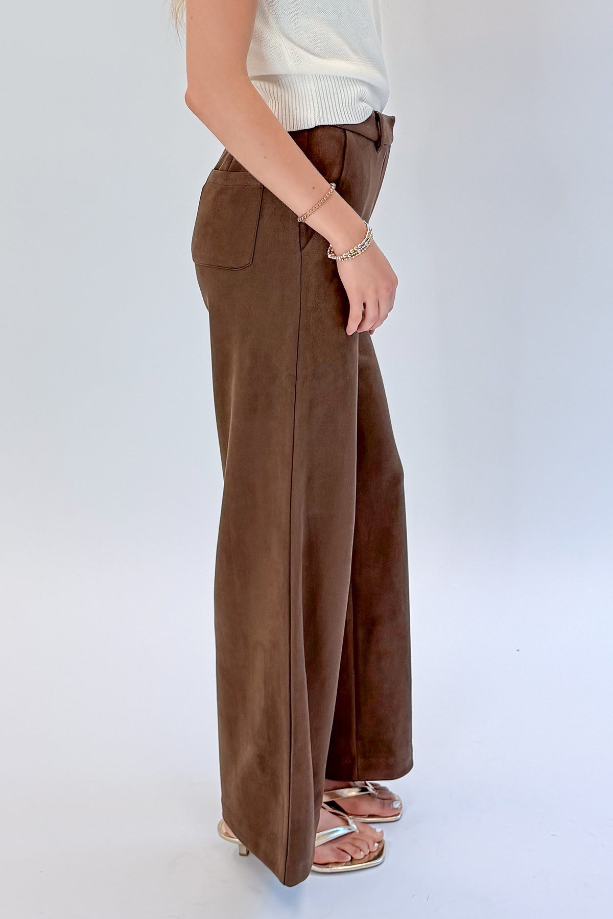 Side view of woman wearing Swagger Boutique’s Chocolate Brown Suede Pants with Living My Best Life White Cap Sleeve Sweater by PINCH against white studio background.