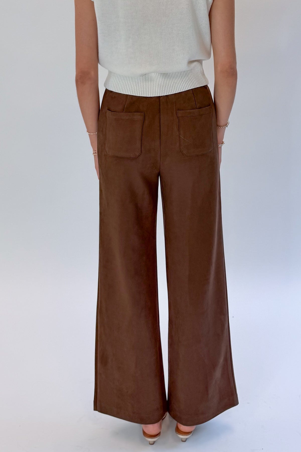 Back view of woman wearing Swagger Boutique’s Chocolate Brown Suede Pants with Living My Best Life White Cap Sleeve Sweater by PINCH against white studio background.