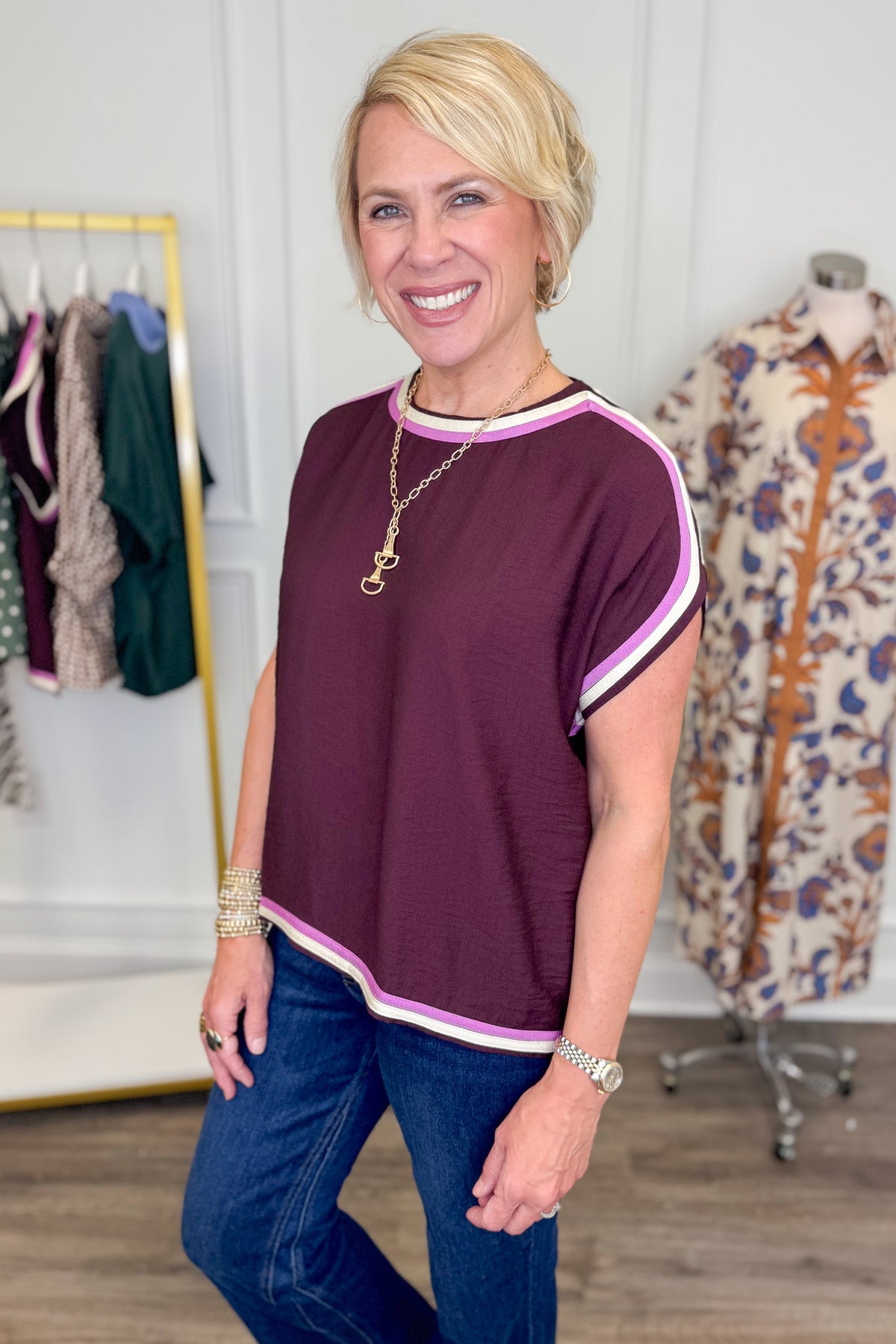 A smiling blonde woman wearing a short-sleeve crewneck top in chocolate with pink and white striped trim, paired with blue jeans, standing in a boutique setting.