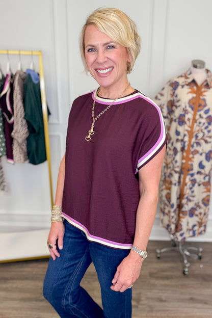 A smiling blonde woman wearing a short-sleeve crewneck top in chocolate with pink and white striped trim, paired with blue jeans, standing in a boutique setting.