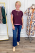 A woman modeling a short-sleeve crewneck top in chocolate with light pink trim, paired with dark blue flared jeans and beige ankle boots, standing in a boutique setting.