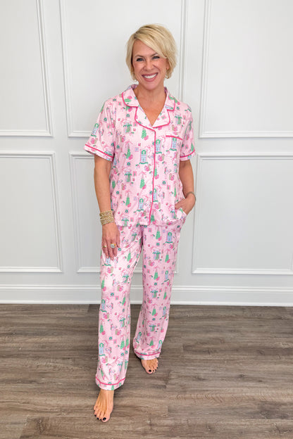 Model wearing the Christmas Cottage Charlotte Pajama Set in pink, featuring a festive Christmas cottage print with green trees, holiday bows, and presents. The set includes a short-sleeve button-down top with contrast piping and matching long pajama pants.