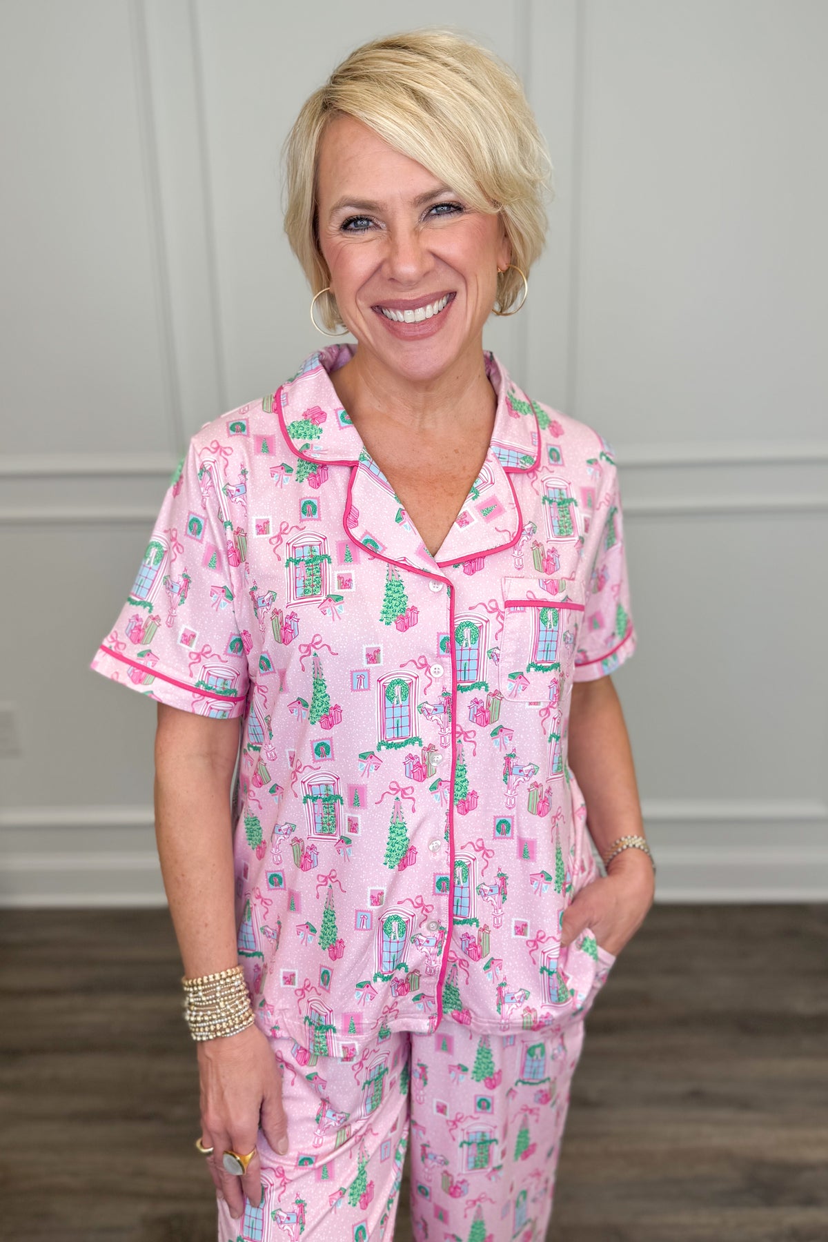 Model wearing the Christmas Cottage Charlotte Pajama Set in pink, featuring a festive Christmas cottage print with green trees, holiday bows, and presents. The set includes a short-sleeve button-down top with contrast piping and matching long pajama pants.