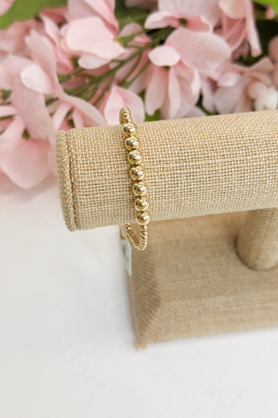 Gold bracelet on a burlap stand with pink flowers in the background