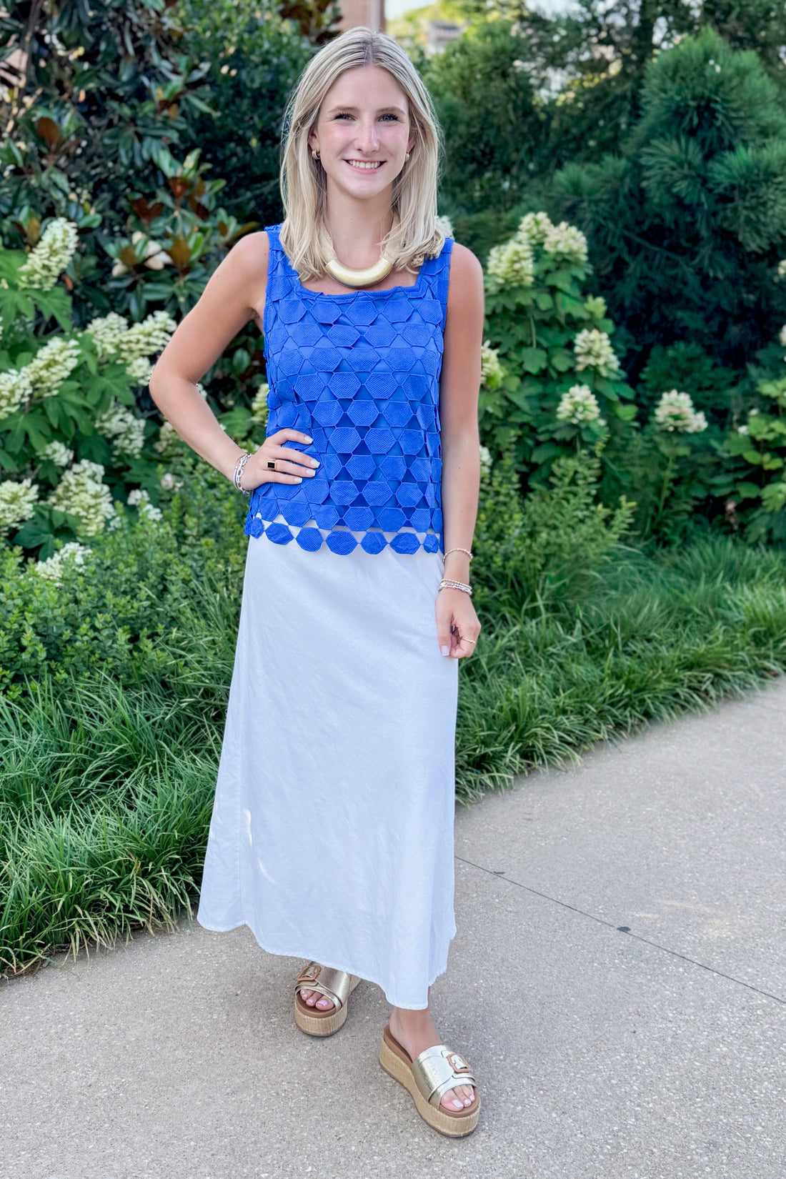 Front view of woman wearing Swagger Boutique’s Cobalt Sleeveless Lace Tank with white skirt standing outside by trees.
