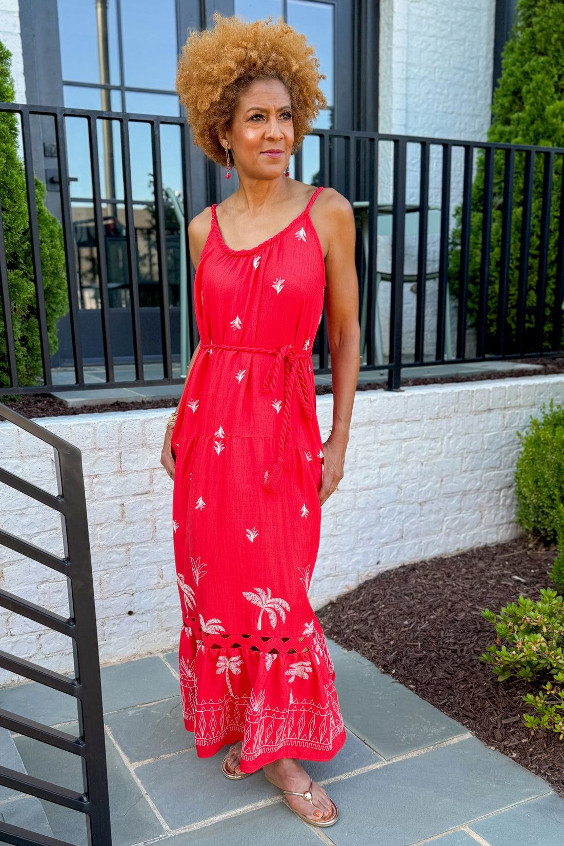 Front view of woman wearing Swagger Boutique’s Coral Palm Embroidered Maxi Dress with sandals standing outside a house.