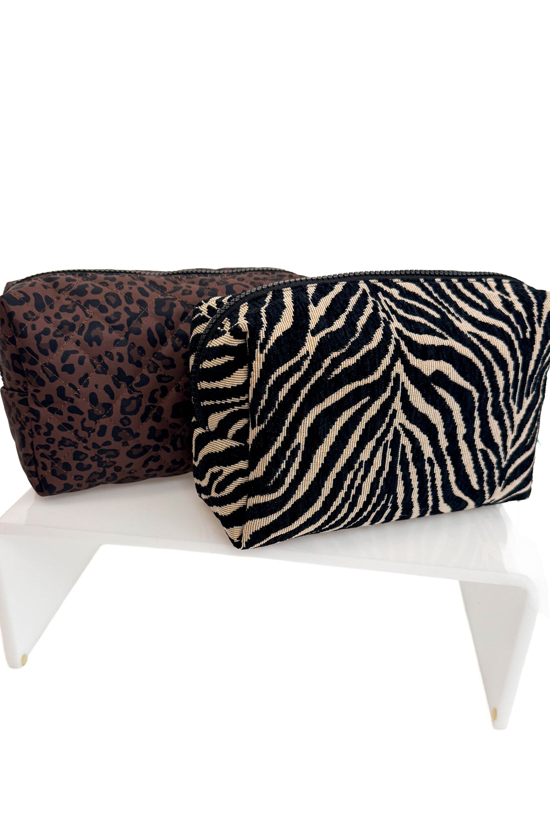 Two zippered cosmetic pouches displayed on a white stand against a white background. The front pouch has a black and beige zebra print, and the back pouch has a dark brown and black leopard print.
