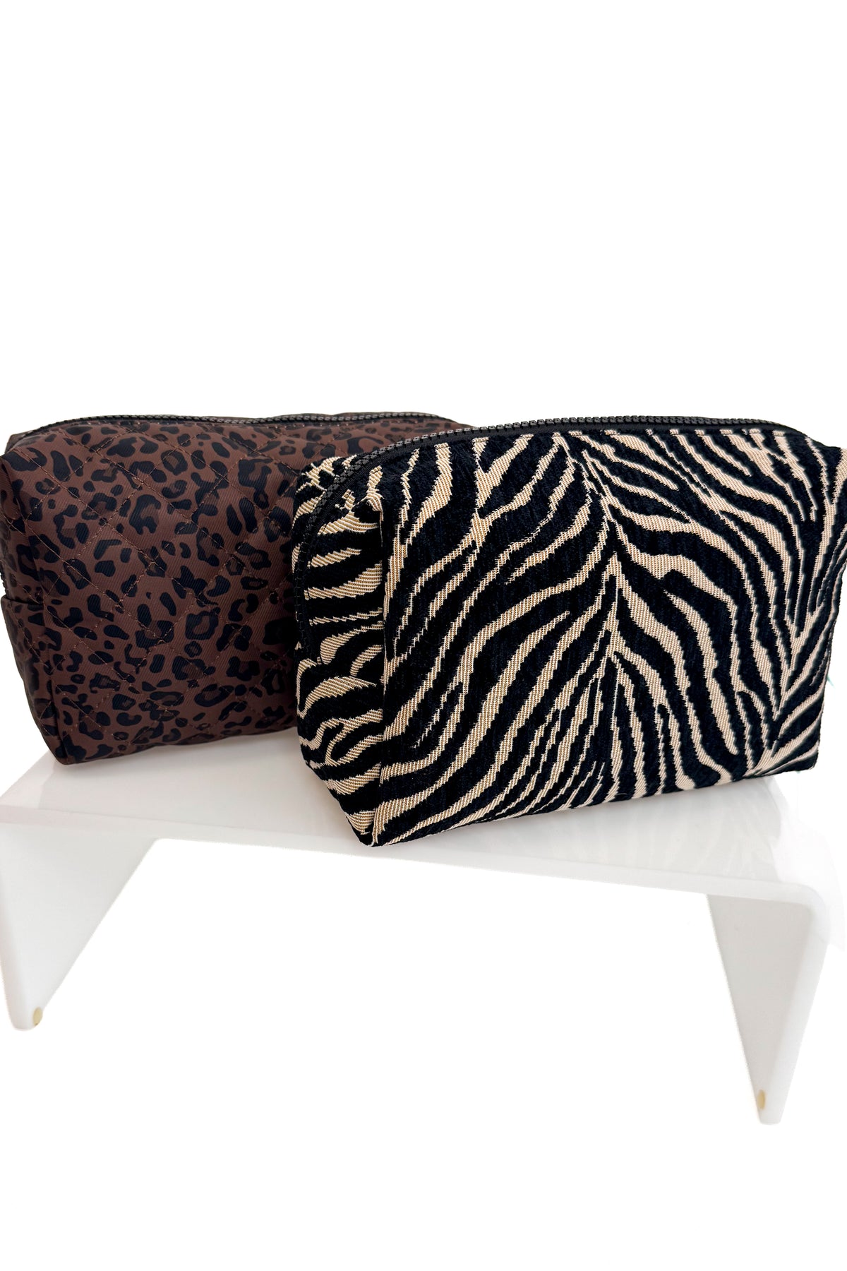 Two zippered cosmetic pouches displayed on a white stand against a white background. The front pouch has a black and beige zebra print, and the back pouch has a dark brown and black leopard print.