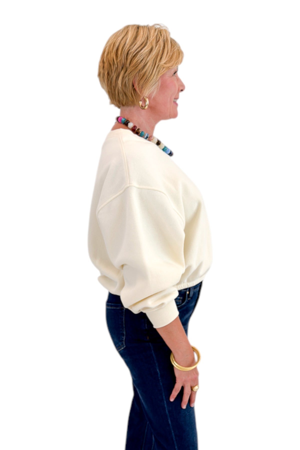 Side view of woman wearing Swagger Boutique’s Cream Elastic Hem Sweatshirt with jeans against white studio background.