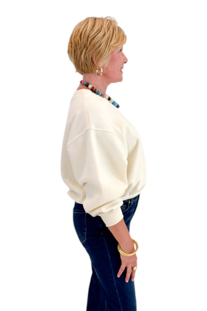 Side view of woman wearing Swagger Boutique’s Cream Elastic Hem Sweatshirt with jeans against white studio background.