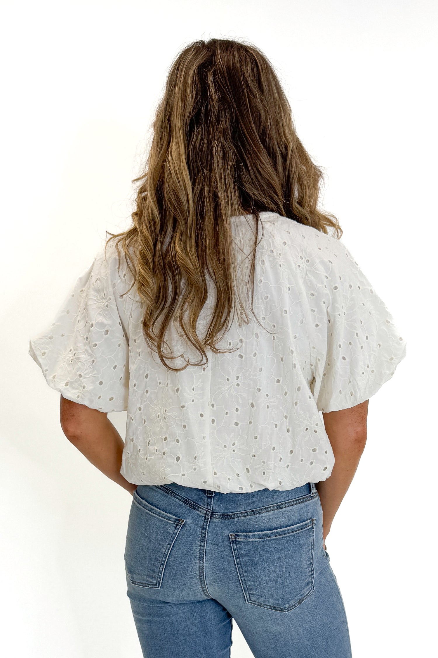 Back view of woman wearing Swagger Boutique’s Cream Eyelet Bubble Hem Top with jeans against white studio background.