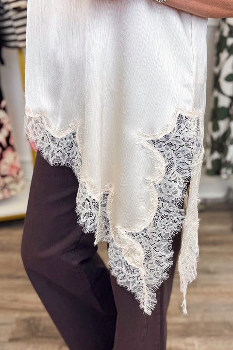 Side detail view of woman wearing Swagger Boutique’s Cream Lace Asymmetrical Tank with jeans with clothing hanging in the background.