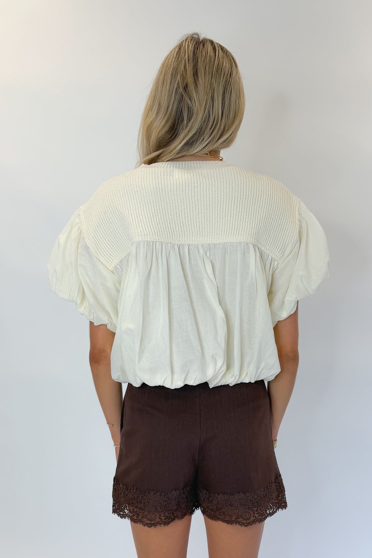 Back view of woman wearing Swagger Boutique’s Brown Lace Lined Denim Skort with Cream Mixed Material Bubble Hem Top against white studio background.