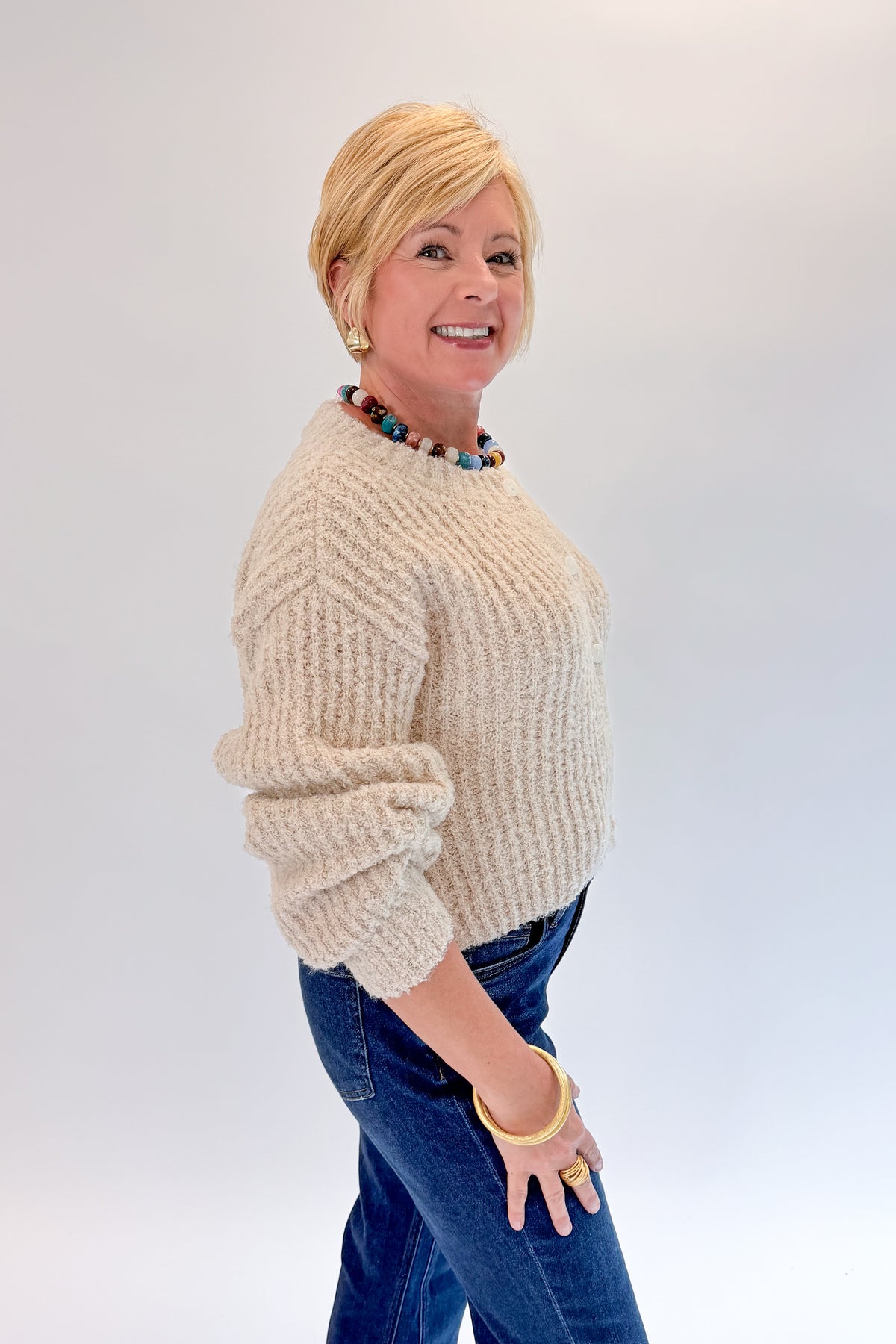 Side view of woman wearing Swagger Boutique’s Cream Ribbed Button Up Cardigan with jeans against white studio background.