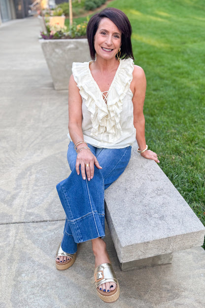 Front view of woman wearing Swagger Boutique’s Cream Sleeveless Ruffle Top with jeans sitting outside. 