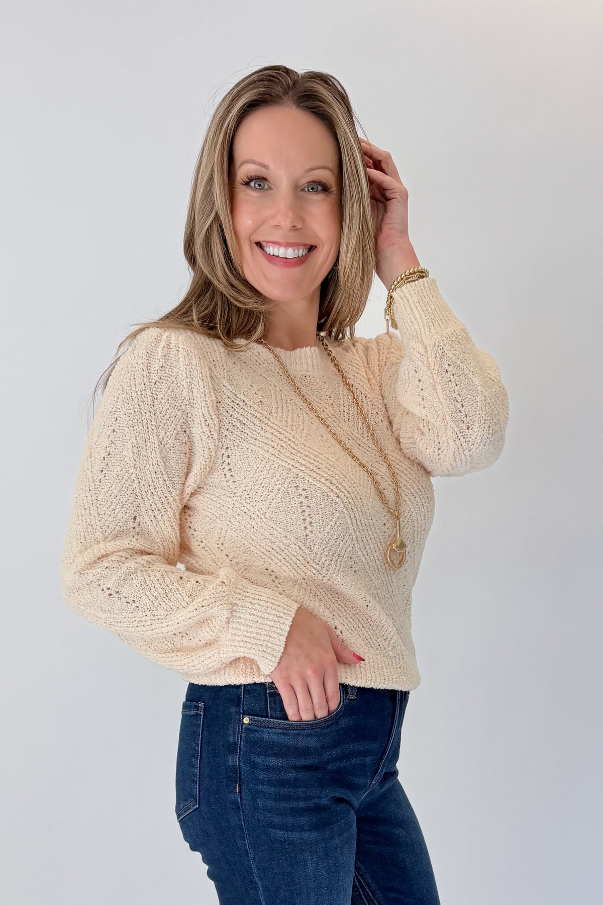 Side view of woman wearing Swagger Boutique’s Cream Textured Crew Neck Sweater with jeans against white studio background.