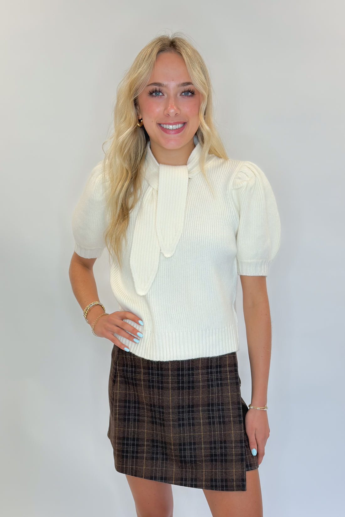 Front view of woman wearing Swagger Boutique’s Cream Tie Neck Short Sleeve Sweater by Entro with Brown Plaid Mini Skirt against white studio background.