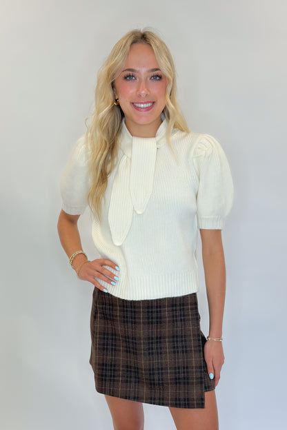Front view of woman wearing Swagger Boutique’s Cream Tie Neck Short Sleeve Sweater by Entro with Brown Plaid Mini Skirt against white studio background.