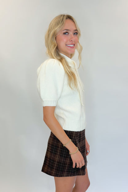 Side view of woman wearing Swagger Boutique’s Cream Tie Neck Short Sleeve Sweater by Entro with Brown Plaid Mini Skirt against white studio background.