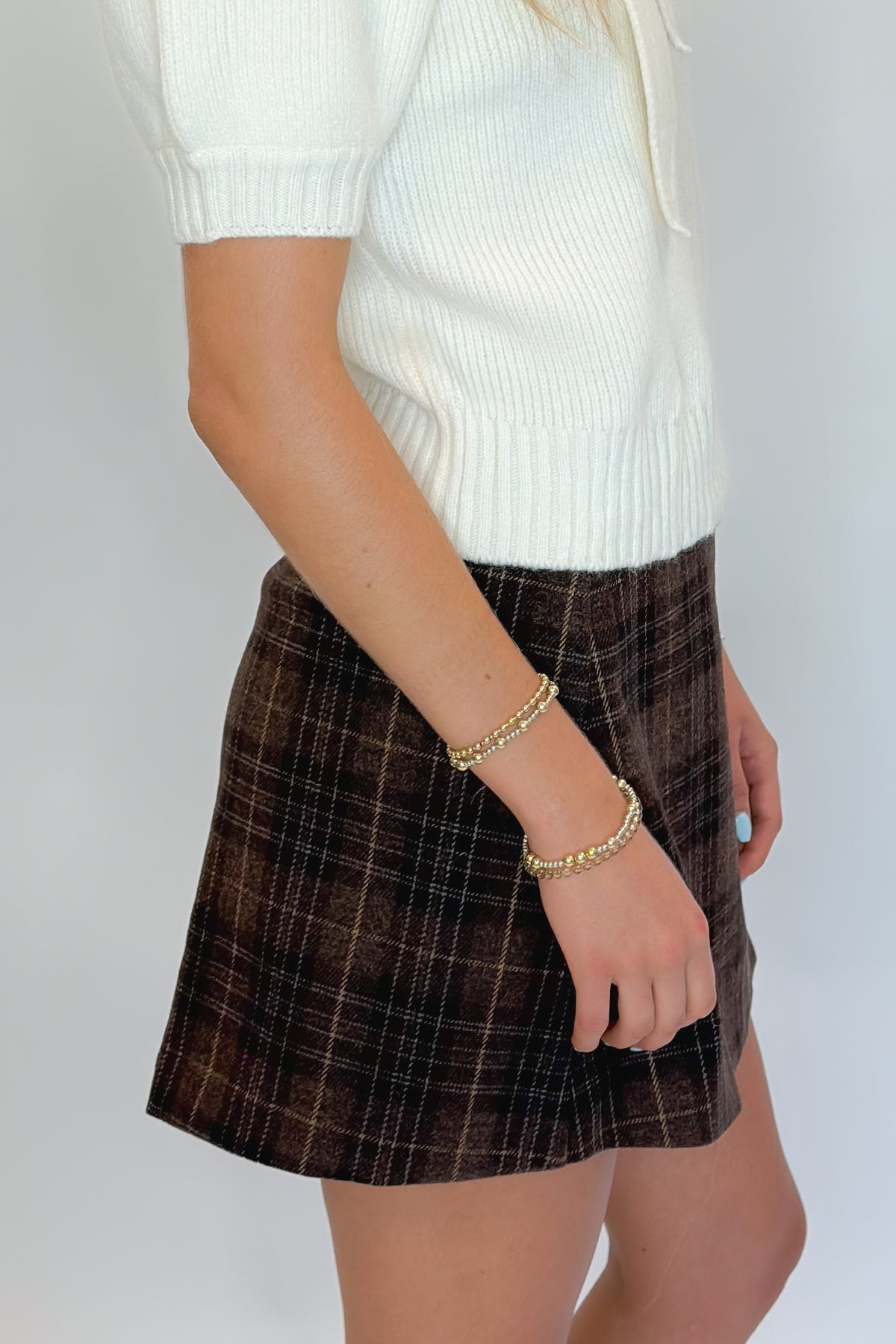 Side view of woman wearing Swagger Boutique’s Cream Tie Neck Short Sleeve Sweater by Entro with Brown Plaid Mini Skirt against white studio background.